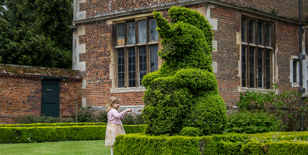 Young girl wearing pink dress in front of large shrubbery cut in the shape of a unicorn by a stately house