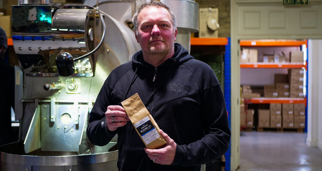 White man wearing black fleece holding bag of coffee in warehouse enivornment
